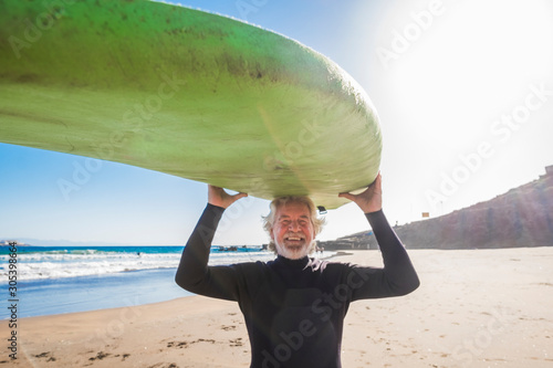 happy senior with surftable on his head is smiling and laughing - old and mature man having fun surfing with a black wetsuits - active retired adult doing activity alone