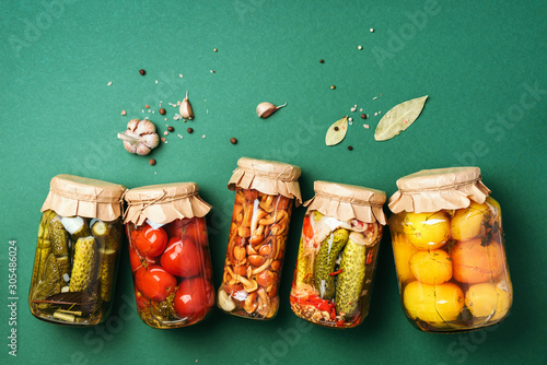 Canned and preserved vegetables in glass jars over green background. Top view. Flat lay. Copy space.