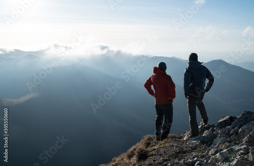 Two men standing standing with trekking poles on cliff edge and looking at sunset rays over the clouds. Successful summit concept image.