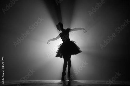 Professional ballerina dancing ballet in spotlights smoke on big stage. Beautiful young girl wearing black tutu dress on floodlights background. Black and white.