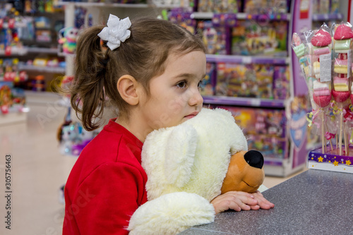 The girl is sad, holding a large polar bear and stands in the store among children's toys. Children's emotions, waiting for gifts and purchases.