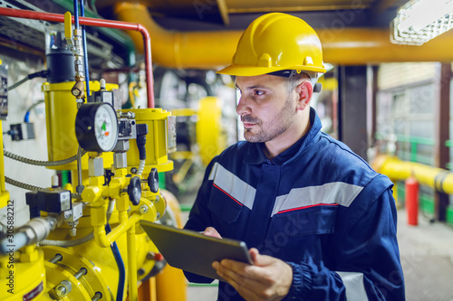 Serious caucasian unshaven worker in protective uniform and with hardhat using tablet for checking temperature in pipes. Factory interior.