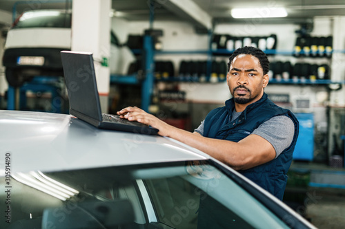 Young black auto mechanic using computer and looking at camera in a workshop.
