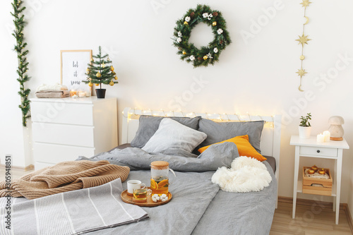 Christmas cozy winter home decor. New year interior decorations. Bed with grey linen, blanket, pillows, plaid, christmas tree, wreath, led garland light, citrus tea on tray. White stylish bedroom.