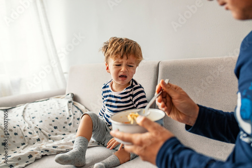 Unhappy child refusing to eat at home with white background while father is trying to deceive the toddler by tricks. Upset unhappy child refuse to eat healthy food