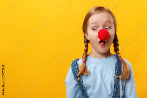 Funny child clown with a red nose. Cheerful little girl on a yellow background. April 1st Fool's Day. Copy space