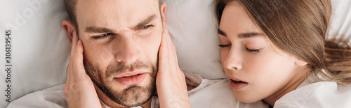 top view of exhausted man plugging ears with hands while lying in bed near snoring wife, panoramic shot