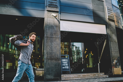 Portrait of bearded hipster guy holding sport bag on back and strolling near shopping center in modern city.Confident young man dressed in casual wear walking in urban setting and going to gym