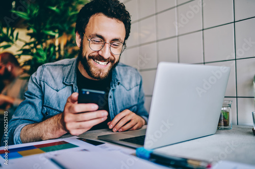 Positive caucasian hipster guy in eyewear laughing at content from social networks using mobile phone for browsing, cheerful male freelancer having fun during remote job chatting on cellular.