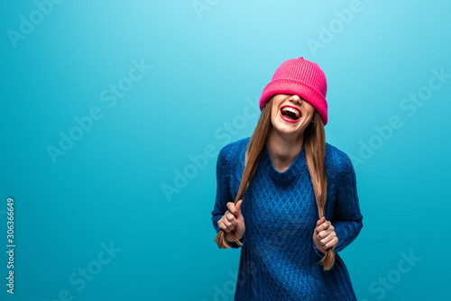 funny laughing woman in knitted sweater with pink hat on eyes, isolated on blue