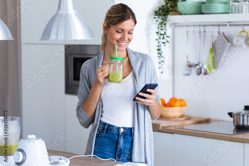 Pretty young woman using her mobile phone while drinking detox juice in the kitchen at home.