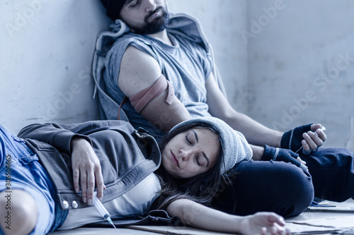 Homeless young man and woman are lying on cardboard on floor in abandoned house. Guy and girl addicts got overdose injections of heroin and getting high. Drug addiction and street life concept.