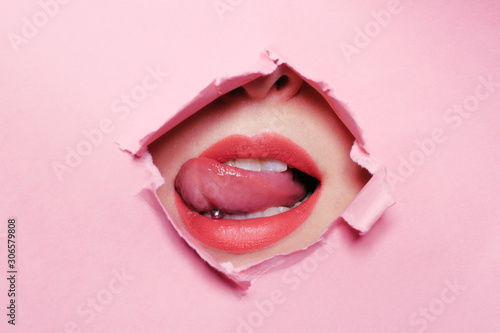 Girl bites her tongue, tongue piercing. Big beautiful lips, piercing parlor. Beautiful teeth, dentistry. Pink background, girl's face through torn cardboard.
