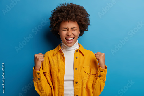 Happy dark skinned woman enjoys moment of success, celebrates victory or great result, feels joyful, gains important goal or achievement, makes yes gesture, dressed in bright clothes, models indoor