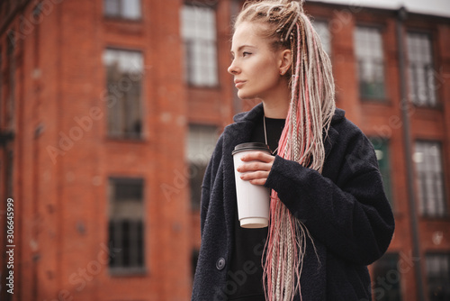 Stylish beautiful young blonde girl with pink dreadlocks with a white glass of take-away coffee, closeup portrait, beauty and fashion concept, trends of millennials and hipsters,