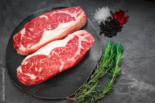 Raw striploin beef steak with rosemary, salt and pepper on round slate on stone table. Top view