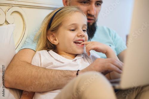 Close up of a lovely little blonde girl biting her finger smiling while her father is choding cartoons on his laptop in the bed .