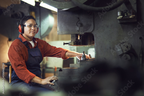 Portrait of smiling African-American woman looking at camera while operating machine units at industrial factory, copy space