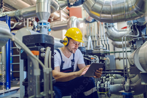 Serious caucasian handsome worker in overall, with hardhat and antiphons crouching and using tablet. Factory interior.