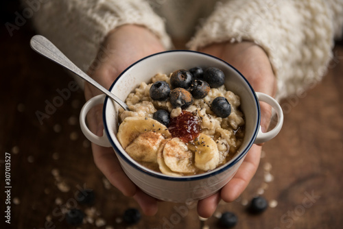 Woman hands offering a vegan porridge with oat flakes, banana, blueberries, chia, cinnamon, maple syrup and strawberry jam. Horizontal. Top view.