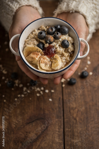 Woman hands holding a healthy porridge with oat flakes, banana, blueberries, chia, cinnamon, maple syrup and strawberry jam. Rustic wooden table in the background. Vertical top view with copyspace