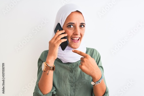 Young Arab woman wearing hijab talking on smartphone over isolated background very happy pointing with hand and finger