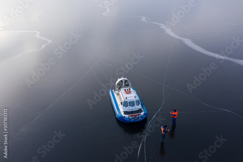 The river is covered with thin ice. On the icy surface is a hovercraft next to the boat are coast guard rescuers. The view from the top.