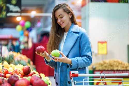 Portrait of attractive woman buyer with cart in the grocery shop during choosing and buying fresh apples at fruit department