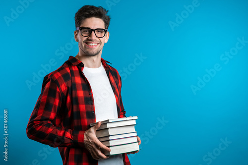 European student in red plaid shirt on blue background in studio holds stack of university books from library. Guy smiles, he is happy to graduate.