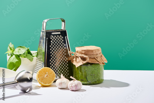 pesto sauce in jar near ingredients, blender and grater on white table isolated on green