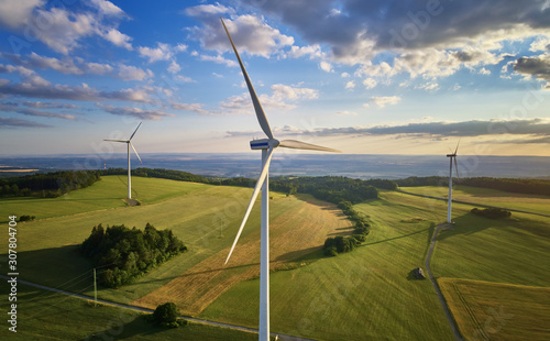 Aerial view of wind turbine farm. Wind power plants in green landscape against sunset sky with clouds. Aerial, drone inspection of wind turbine.