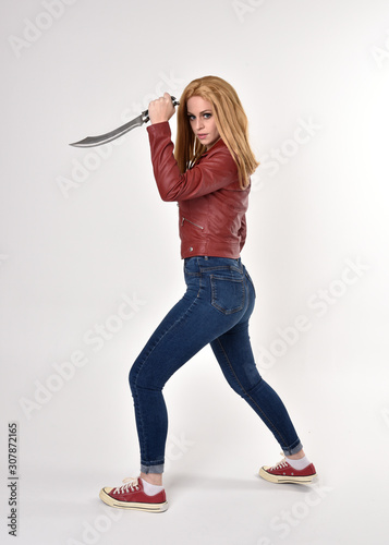 Full length portrait of a pretty blonde girl wearing red leather jacket denim jeans and sneakers. Standing pose, holding a dagger, on a studio background.