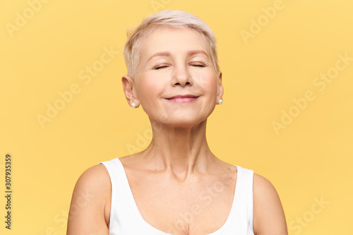 Fashionable beautiful retired Caucasian woman with pixie hairdo wearing casual clothes posing in studio keeping eyes closed and smiling with pleasure and enjoyment, listening to good music or dreaming
