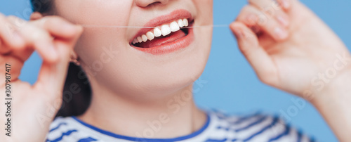 Happy smiling woman with white perfect teeth flossing, dental floss on blue background, clode up photo.