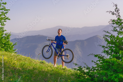 Tired cyclist is wiping his sweat off his face while pushing his bicycle uphill on a dirt road in a mountain.