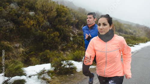 Couple of motivated athletes running under cold winter snow storm on mountain road.