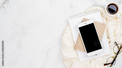 Feminine home office desk concept. White marble background with smartphone, paper notepad, glasses, cup of coffee. Top view, flat lay, copy space.