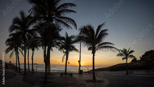 Panorama of a sunrise on the Arpoador boulevard with the Devils beach and silhouetted palm trees in Rio de Janeiro against a clear orange and blue sky