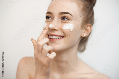 Portrait of a charming young female with red hair and freckles isolated on white applying a anti age cream on her face and nose smiling.
