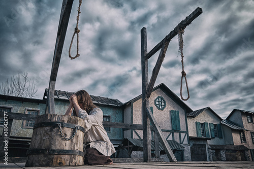 A woman in medieval dress prays against a cloudy dramatic sky. Place of execution with gallows and scaffold on the background of old houses
