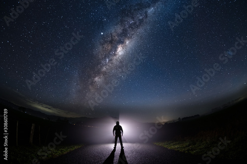 A mysterious man standing in the middle of the road looking into bright light with milky way starry night sky.