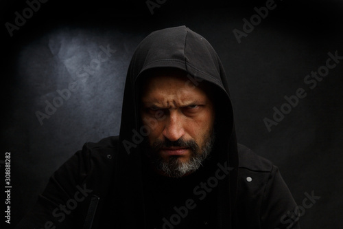 Portrait of a bald man with a beard in a black hood on a dirty gray background.