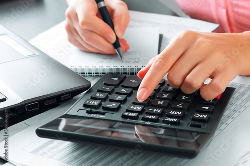 Female accountant in the office uses a calculator and writes data in a notebook. Profit analysis, taxes and payments calculations, preparation of financial statements concept