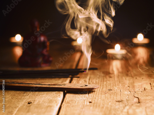 Smoking incense stick in the foreground. On background Small statue of Buddha with incense sticks and burning candles