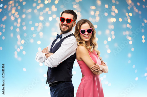 valentines day, love and people concept - happy couple in heart-shaped sunglasses over holiday lights on blue background