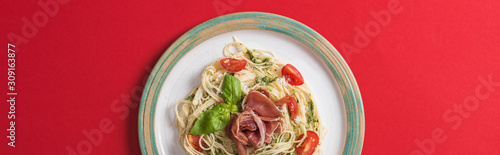 top view of cooked Pappardelle with tomatoes, basil and prosciutto on plate on red background, panoramic shot