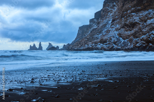 Landscape with the beach of black sand, dramatic sky and the sea stacks in the background at twilight in winter, Reynisdrangar (near Vik), Iceland.