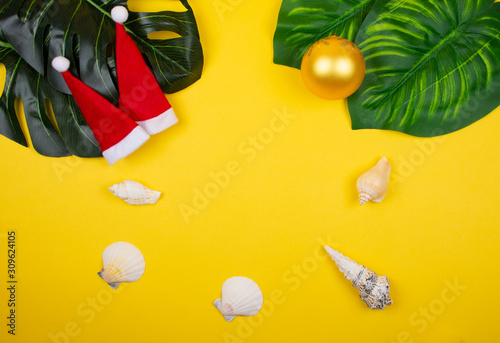 Two small Santa hats, tropical leaves, seashells and a Christmas ball on the bright yellow background as the tropical Christmas concept (copy space in the center)