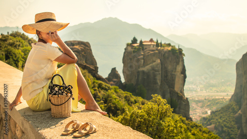 Young woman with white dress and large hat sitting in front of greece meteor mountains, monastery and village in the background