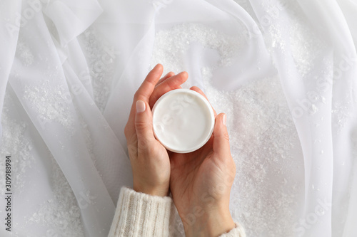 Woman holding jar of winter cream for skin, decorative snow on white fabric background, top view. Space for text